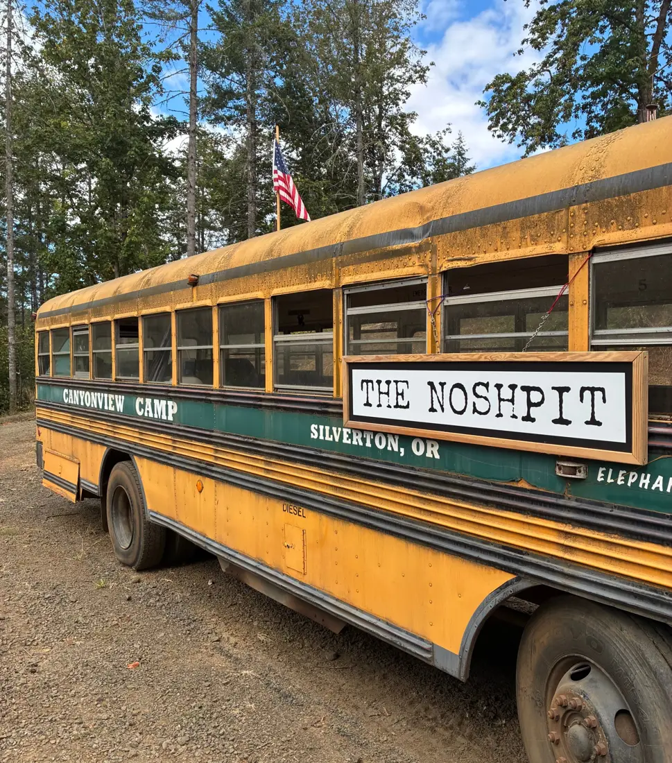 The bus with a wooden-framed NoshPit sign hung on its side, parked on gravel under tall trees